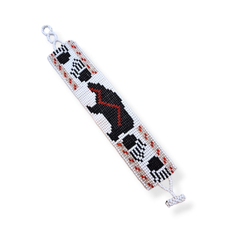 May include: White beaded bracelet with a black bear design and red accents. The bracelet has a clasp closure.