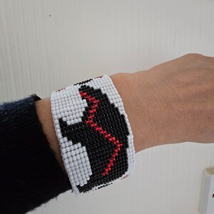 May include: A white beaded bracelet with black and red beads forming a geometric pattern. The bracelet is worn on a person's wrist.