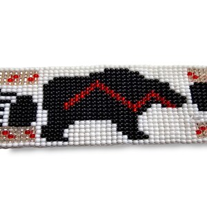 May include: A beaded bracelet with a black and white bear design. The bear is outlined in red beads. The bracelet is made with white, black, and red beads.