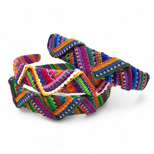 May include: Two colourful woven headbands. The headbands display a vibrant mix of colours including blue, green, red, and purple. The fabric is intricately woven with a zig-zag pattern. The headbands are likely made of fabric and are suitable for various occasions.