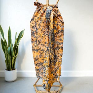 Peut inclure: Un paréo à motifs orange et noir est présenté sur un support en bambou. Le tissu présente un motif détaillé avec des formes florales et abstraites. La texture et les couleurs vives du paréo sont mises en valeur par son drapé. Une plante en pot est en arrière-plan.