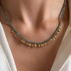 May include: A delicate necklace featuring small, gold-colored coin pendants suspended from a strand of turquoise-colored beads. The necklace is worn against a white collared shirt, highlighting the jewelry's design.