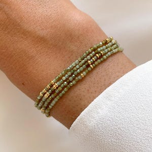 Peut inclure: Quatre bracelets de perles. Les bracelets sont composés de petites perles vert clair et de perles dorées. Ils sont portés sur un poignet, sur un fond blanc.