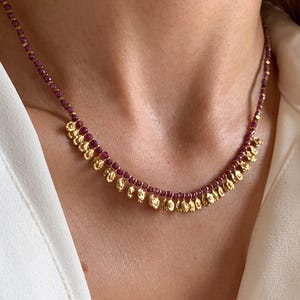 Può includere: Una delicata collana con piccole perline rotonde di colore rosso scuro infilate insieme. La collana è impreziosita da ciondoli dorati a forma di goccia che pendono sul davanti. La collana è esposta su una camicia bianca.