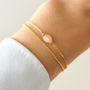 Könnte beinhalten: Ein zartes goldenes Kettenarmband mit einem einzigen rosa ovalen Edelstein-Anhänger. Das Armband ist mit einer zweiten, dünneren goldenen Kette geschichtet.