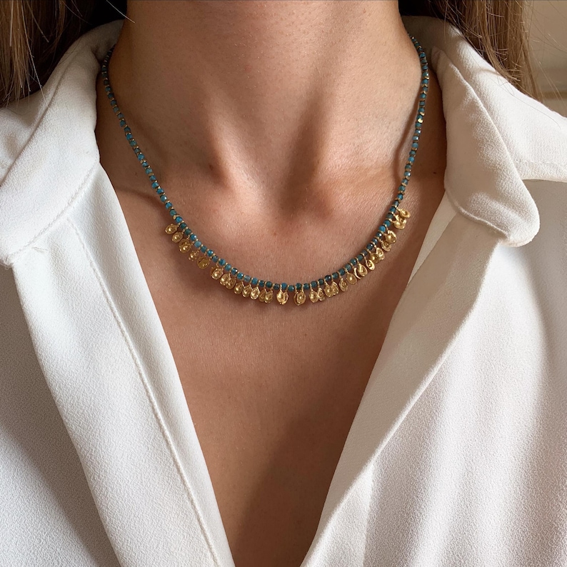 May include: A delicate necklace featuring small, gold-colored coin pendants suspended from a strand of turquoise-colored beads. The necklace is worn against a white collared shirt, highlighting the jewelry's design.