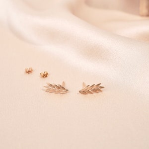 May include: A pair of gold leaf earrings on a light beige fabric background.