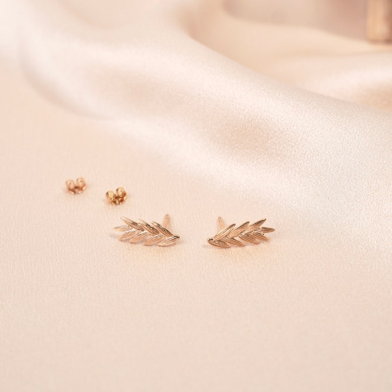 May include: A pair of gold leaf earrings on a light beige fabric background.