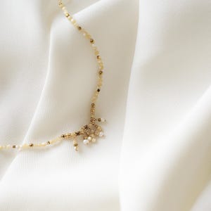 May include: Delicate necklace featuring small, light-colored beads and gold accents. The necklace has a cluster of small, dangling charms at the center. The jewelry is displayed on a white, textured fabric.