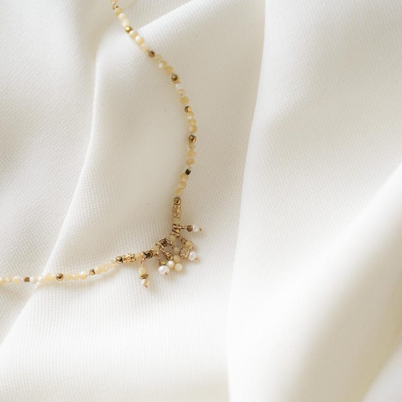 May include: Delicate necklace featuring small, light-colored beads and gold accents. The necklace has a cluster of small, dangling charms at the center. The jewelry is displayed on a white, textured fabric.