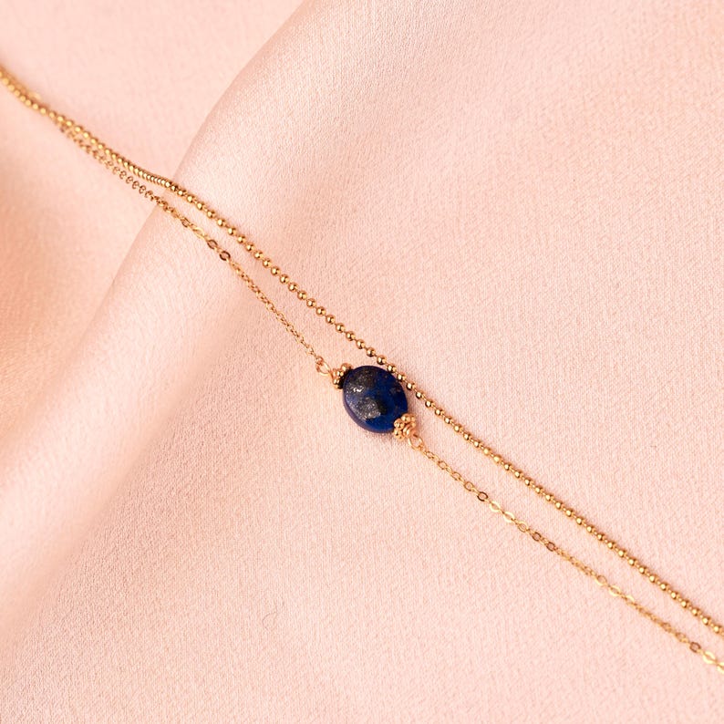 May include: A delicate gold chain necklace with a single, oval-shaped lapis lazuli stone pendant. The necklace is layered with two chains of different thicknesses.