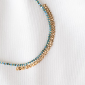 May include: A gold-toned necklace with turquoise beads and small, dangling gold-colored pendants. The necklace is set against a white background, highlighting its delicate design and intricate details. The necklace is a piece of jewelry.