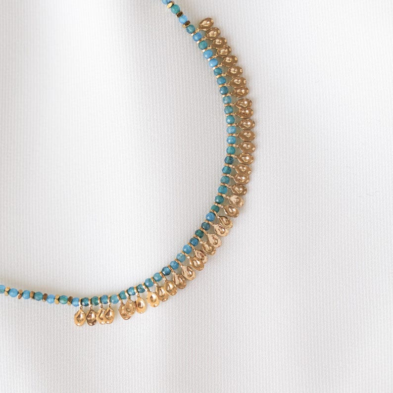 May include: A gold-toned necklace with turquoise beads and small, dangling gold-colored pendants. The necklace is set against a white background, highlighting its delicate design and intricate details. The necklace is a piece of jewelry.