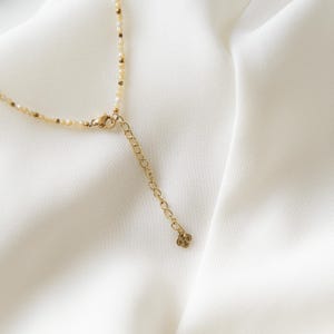May include: Delicate necklace featuring small, light-colored beads and gold-toned accents. The necklace has a gold clasp and an adjustable chain. The background is a white, textured fabric.