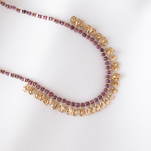 May include: A delicate necklace featuring small, round, purple beads interspersed with gold-colored, square beads. The necklace has a curved design with gold-colored, teardrop-shaped pendants hanging from the bottom. The background is a plain white.
