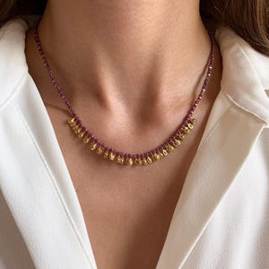 May include: A delicate necklace featuring small, faceted, deep red beads strung together. The necklace has a series of gold-colored, teardrop-shaped pendants hanging from the bottom. The necklace is worn against a white shirt.