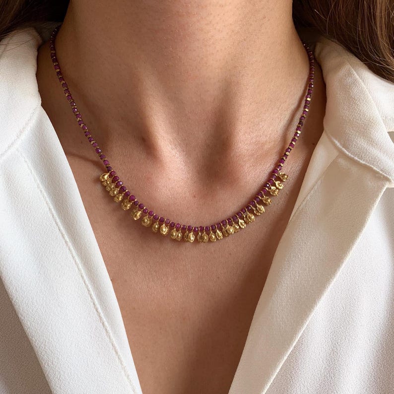 May include: A delicate necklace featuring small, faceted, deep red beads strung together. The necklace has a series of gold-colored, teardrop-shaped pendants hanging from the bottom. The necklace is worn against a white shirt.
