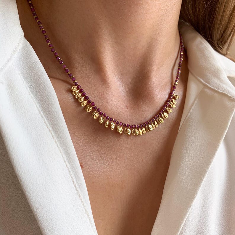 May include: A delicate necklace featuring small, round, deep red beads strung together. The necklace has gold-colored, teardrop-shaped pendants hanging from the bottom. The necklace is worn against a white shirt.