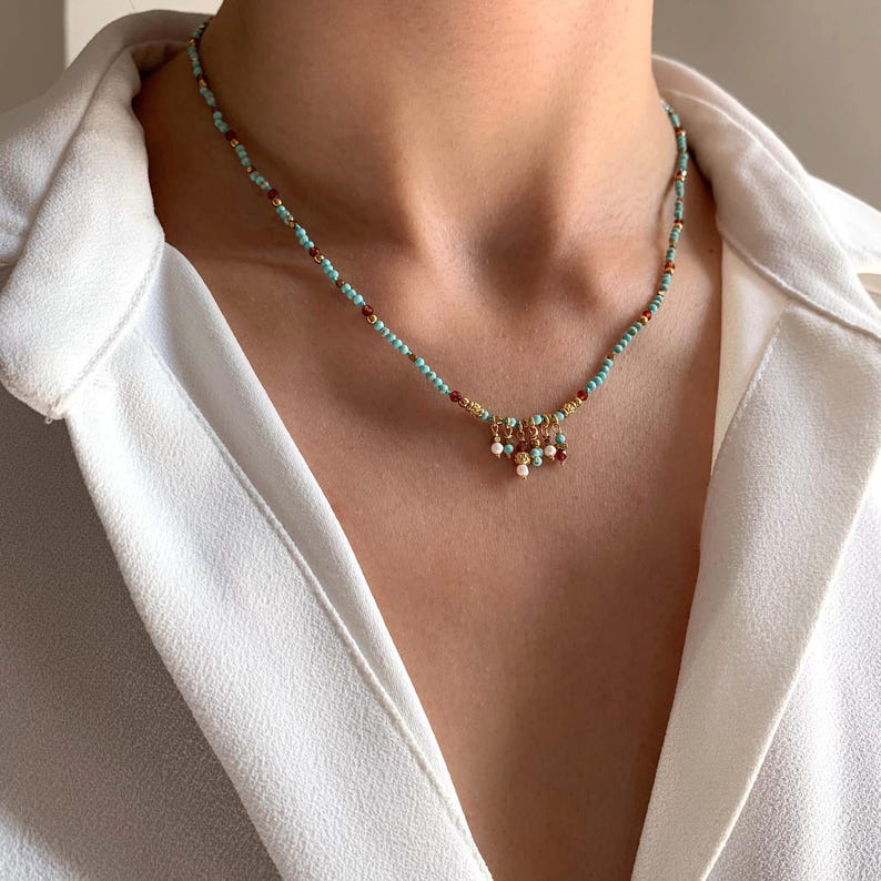May include: A delicate necklace featuring small turquoise beads interspersed with red and gold accents. The necklace has a gold pendant with dangling white, red, and gold beads. The necklace is worn against a white shirt.