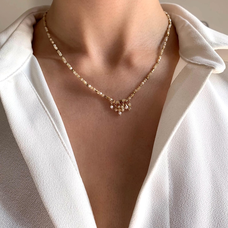 May include: Delicate necklace featuring small, light-colored beads and gold accents. The necklace has a central cluster of dangling beads, creating a layered effect. The jewelry is worn against a white shirt.
