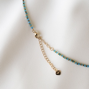 May include: Close-up of a delicate necklace featuring small, light blue beads interspersed with gold-colored accents. The necklace has a gold-toned chain and clasp, with an adjustable extender. The design is simple and elegant.