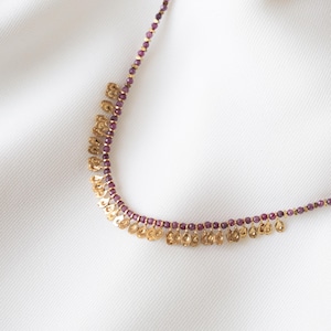 May include: A delicate necklace featuring small, round, purple beads and gold-colored charms. The charms are in a floral or petal-like shape, hanging in a fringe along the bottom of the necklace. The necklace is displayed on a white surface.