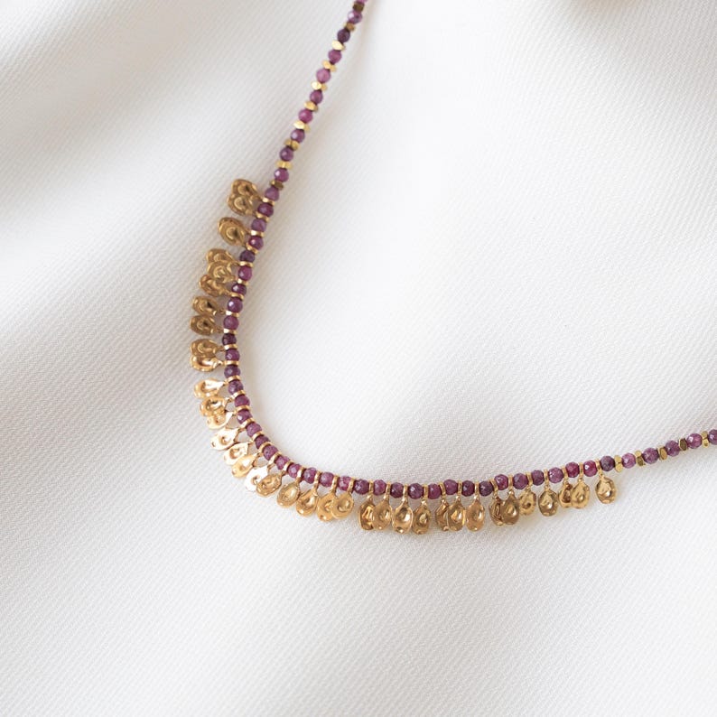 May include: A delicate necklace featuring small, round, purple beads and gold-colored charms. The charms are in a floral or petal-like shape, hanging in a fringe along the bottom of the necklace. The necklace is displayed on a white surface.
