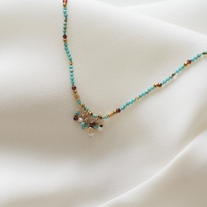 May include: Delicate turquoise beaded necklace with gold accents and small dangling clusters of pearls and red beads. The necklace is displayed on a white, textured surface.