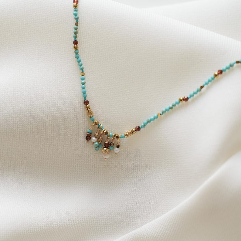 May include: Delicate turquoise beaded necklace with gold accents and small dangling clusters of pearls and red beads. The necklace is displayed on a white, textured surface.