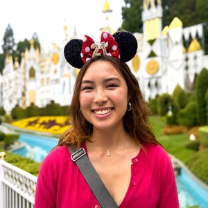May include: A headband featuring two black sequined ears and a central red bow with white polka dots. The bow displays the number 21 in gold glitter. The headband is worn on the head, showcasing the decorative ears and the prominent gold number 21.