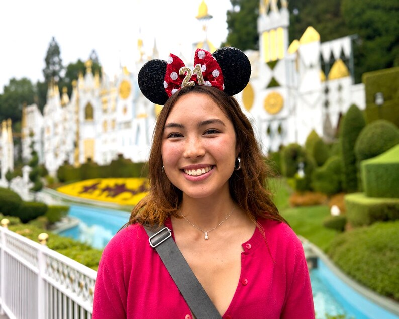 May include: A headband featuring two black sequined ears and a central red bow with white polka dots. The bow displays the number 21 in gold glitter. The headband is worn on the head, showcasing the decorative ears and the prominent gold number 21.