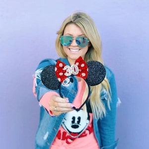 May include: A black headband featuring two round, glittery black ears and a central red bow with white polka dots. The bow is embellished with a silver glittery number 30 centered on the front. The headband is constructed with a thin black frame.
