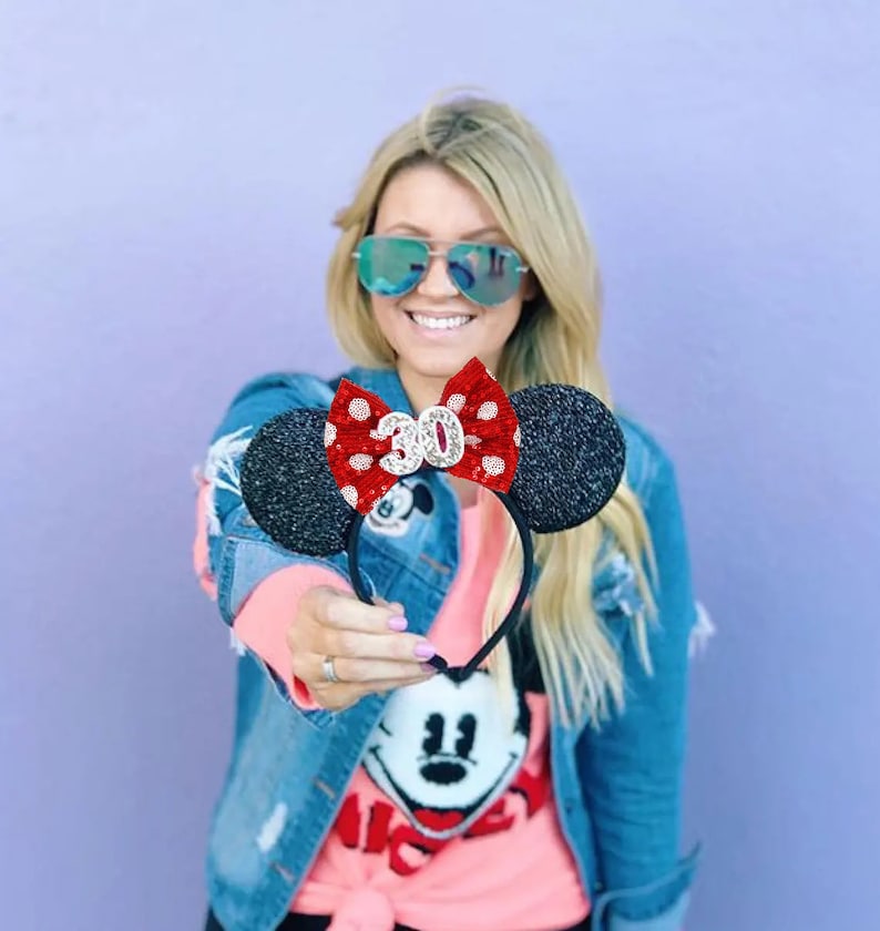 May include: A black headband featuring two round, glittery black ears and a central red bow with white polka dots. The bow is embellished with a silver glittery number 30 centered on the front. The headband is constructed with a thin black frame.