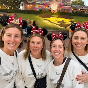 May include: Four white long-sleeve sweatshirts featuring a grey graphic print of a cartoon character and bats, paired with black sequined Minnie Mouse ear headbands that have red and white polka dot bows and a gold 40th anniversary emblem.