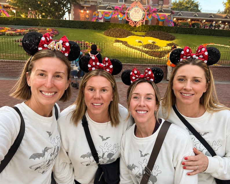May include: Four white long-sleeve sweatshirts featuring a grey graphic print of a cartoon character and bats, paired with black sequined Minnie Mouse ear headbands that have red and white polka dot bows and a gold 40th anniversary emblem.