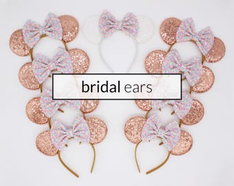 May include: A collection of headbands featuring round ears covered in rose gold sequins and topped with bows in a multi-colored floral pattern. One white headband with a white bow is included. The text "bridal ears" is centered in a black box.