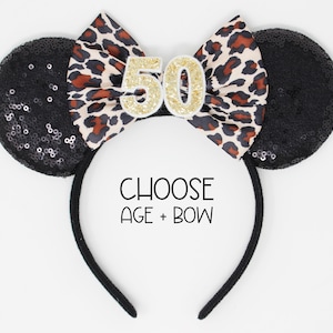 May include: A black headband featuring two round ears covered in black sequins and a central bow with a leopard print pattern. The number 50 in gold glitter is attached to the bow. The text CHOOSE AGE + BOW is printed in black below the bow.