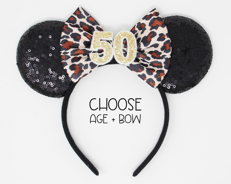 May include: A black headband featuring two round ears covered in black sequins and a central bow with a leopard print pattern. The number 50 in gold glitter is attached to the bow. The text CHOOSE AGE + BOW is printed in black below the bow.