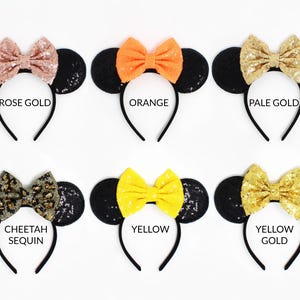 May include: Six headbands with black sequin ears and fabric bows in rose gold, orange, pale gold, cheetah print, yellow, and yellow gold. Each headband features a central bow and round ears, with text labels identifying each color variant below.