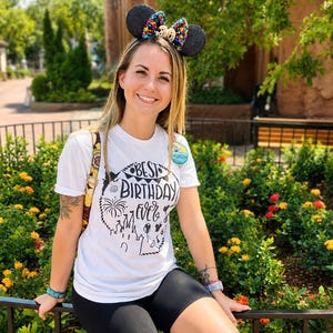 May include: A white short-sleeved t-shirt featuring a black graphic print that reads Best Birthday Ever with illustrations of a castle, fireworks, and balloons. The shirt is paired with black shorts and a black headband with ears and a colorful bow.