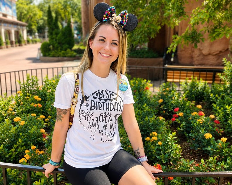 May include: A white short-sleeved t-shirt featuring a black graphic print that reads Best Birthday Ever with illustrations of a castle, fireworks, and balloons. The shirt is paired with black shorts and a black headband with ears and a colorful bow.