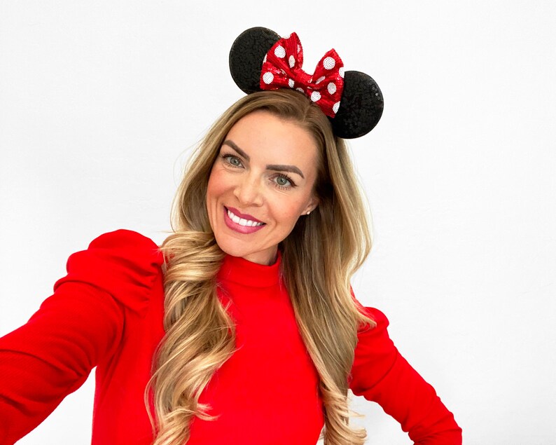May include: A woman with long blonde hair wearing a red mock neck shirt and a black and red polka dot headband with mouse ears.