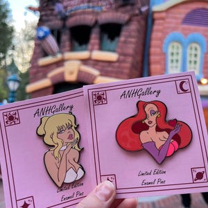 May include: Two pink cards with enamel pins featuring cartoon women. The pins are labeled "ANHGallery" and "Limited Edition Enamel Pins." One pin has a blonde woman blowing a kiss, and the other has a red-haired woman holding a heart.