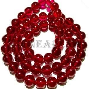 Pode incluir: Um colar de contas redondas vermelho-escuro translúcidas. As contas são enfiadas juntas. Parecem ser de vidro ou material semelhante. Fundo branco.