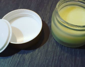 May include: A small glass jar with a gold lid filled with a yellow balm or cream. The jar is sitting on a dark wooden surface. The lid is open and the jar is partially empty.