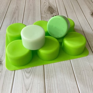 May include: A green silicone mould with six round cavities, filled with five round bars of soap. Two of the soap bars are light green, and three are a darker shade of green.
