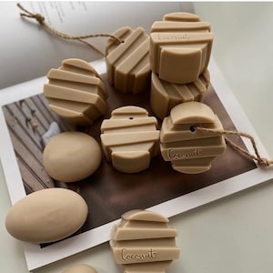 Pode incluir: Uma coleção de sabonetes bege em várias formas, incluindo geométricas e ovais, alguns com a palavra "Coconut" gravada. Os sabonetes estão dispostos numa página de revista, alguns com barbante. A estética geral é neutra e minimalista.
