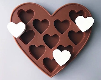 Puede incluir: Un molde de silicona en forma de corazón marrón con 12 cavidades en forma de corazón. Tres objetos en forma de corazón blancos se colocan en el molde.