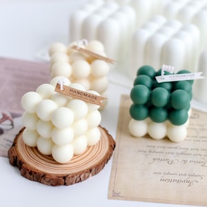 Op de afbeelding: Twee vierkante kaarsen met een bubbelstructuur. Eén kaars is wit en de andere is groen. Beide kaarsen hebben een klein bruin label met de tekst "Handmade". De kaarsen staan op een houten oppervlak.
