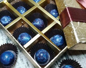 May include: A gold box with a brown ribbon and a lid open to reveal a tray of dark blue and silver chocolates. The chocolates are arranged in a grid pattern in the tray.